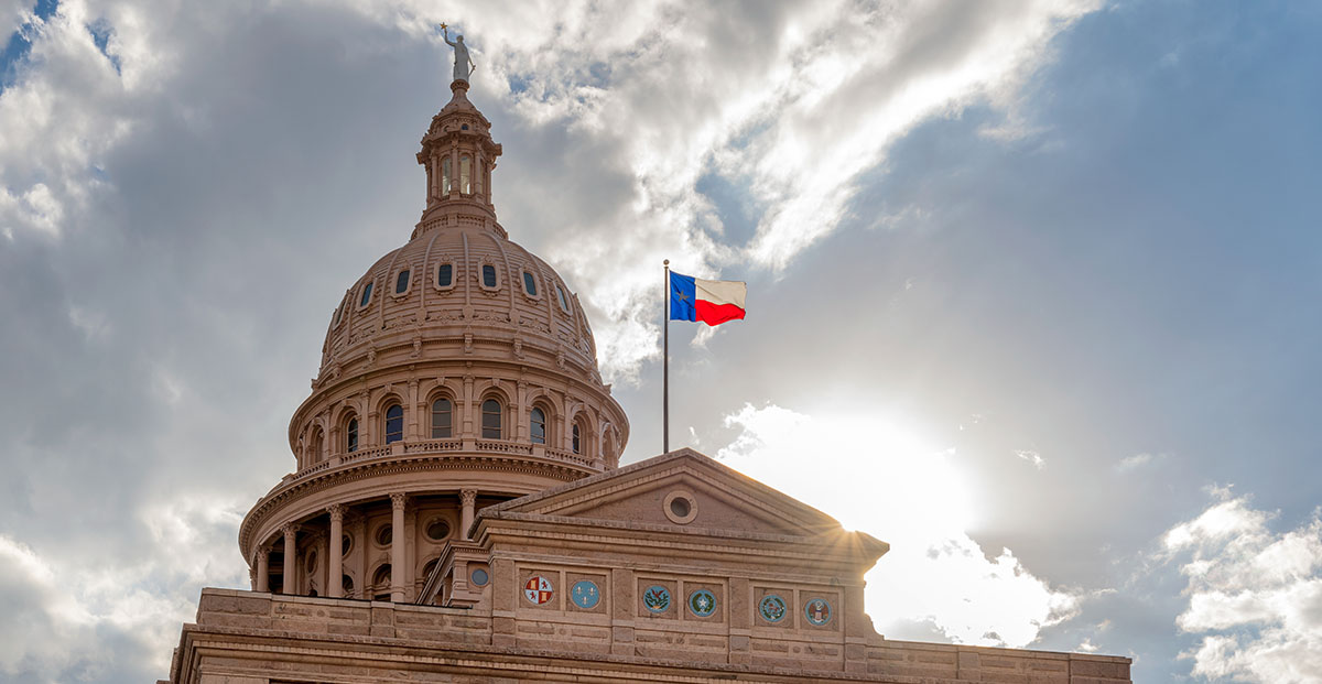 The Texas state flag flies over the State Capitol, against a partially cloudy sky.